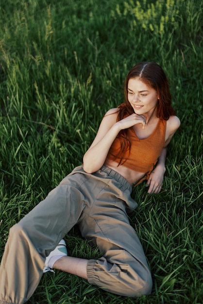 Una mujer tendida en la hierba verde con pantalones verdes y una blusa naranja sonriendo al sol de verano al atardecer en la naturaleza