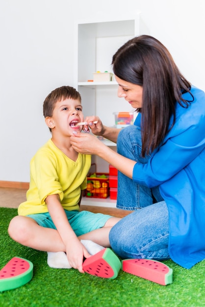 Una mujer terapeuta del habla trata con un niño y realiza un ejercicio para corregir el aparato del habla jugando en el suelo.