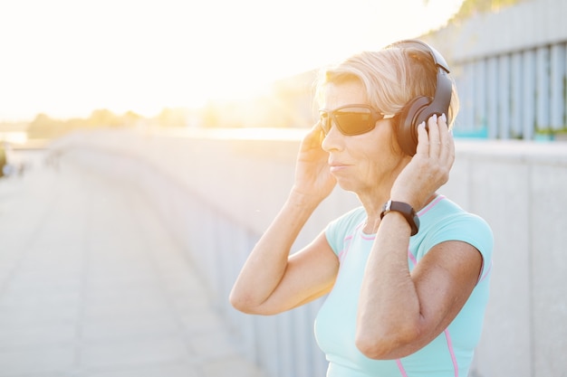 La mujer de la tercera edad activa disfruta de escuchar música después del entrenamiento físico en la ciudad con auriculares y gafas de sol. Reloj inteligente. Mujer fitness deportiva. Estilo de vida saludable. Foto de alta calidad