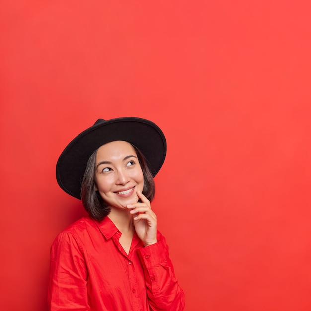 La mujer tiene la expresión de la cara alegre mantiene la mano en la cara enfocada arriba vestida con un atuendo de moda posa en rojo vivo