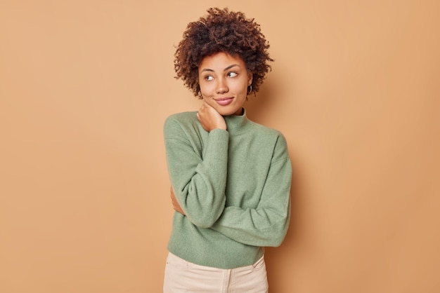 La mujer tiene expresión satisfecha, mantiene la mano en el cuello concentrada, piensa en un momento agradable en la vida, usa un jersey casual aislado en beige, advierte a un amigo