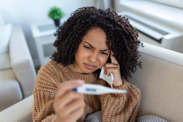 La mujer tiene gripe y está usando un termómetro Enferma con rinitis Mujer con goteo nasal Mujer enferma con gripe acostada en el sofá mirando la temperatura en el termómetro