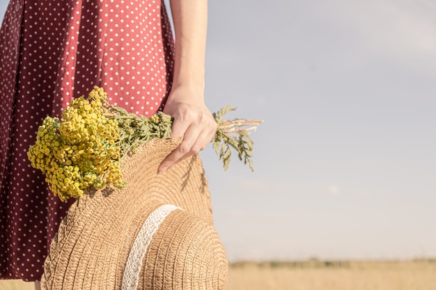 Mujer tiene ramo de flores de campo y un sombrero. Escena rural: vista de primer plano de mujer con vestido de lunares con sombrero de granjero y ramo en la mano