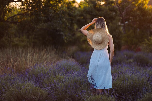 Mujer tierna se encuentra de espaldas con vestido blanco y sombrero entre lavanda e iluminada por rayos de sol