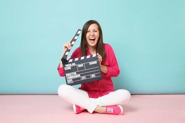 Mujer de tiro de longitud completa en pantalones blancos de camisa rosa sentada en el piso con claqueta de cine