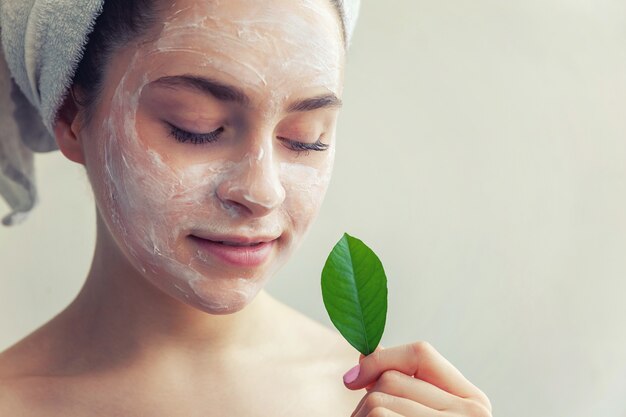 Mujer en toalla en la cabeza con mascarilla nutritiva blanca o crema en la cara y hoja verde en la mano, fondo blanco aislado