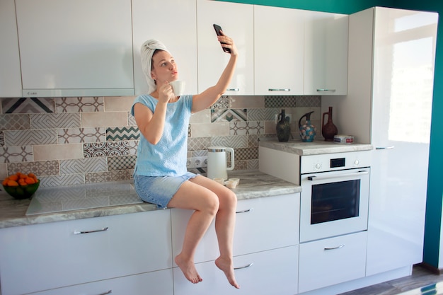 Mujer con una toalla en la cabeza en su cocina desayunando y mirando el teléfono