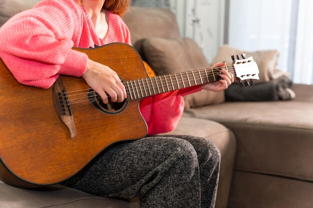 Mujer tocando la guitarra en casa sentada en el sofá de su sala de estar al lado de la venta