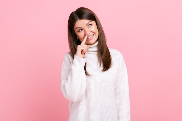 Mujer tocando la nariz mostrando un gesto mentiroso enojado por la falsedad engaño absoluto noticias falsas usando un suéter blanco de estilo casual Foto de estudio interior aislada en fondo rosa