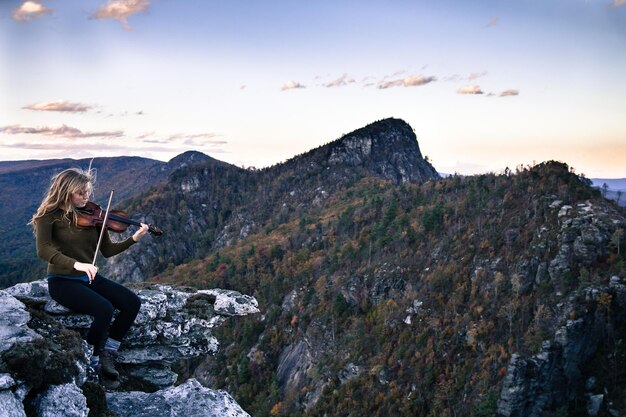 Foto mujer tocando el violín mientras está sentada en una roca contra las montañas