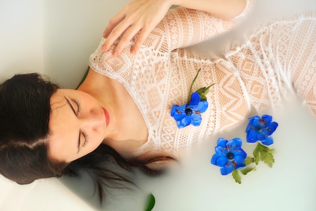 La mujer toma un baño de bienestar lleno de leche, los capullos de flores azules flotan en la superficie y se relajan.