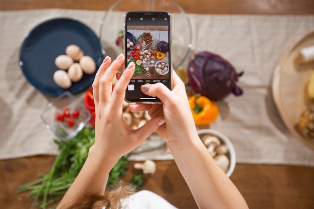 La mujer toma una foto de diferentes verduras y verduras hermosas con un teléfono móvil en su cocina. Fotografía de alimentos en teléfonos inteligentes. Alimentación saludable, comida vegetariana, dietas. para pérdida de peso y desintoxicación.