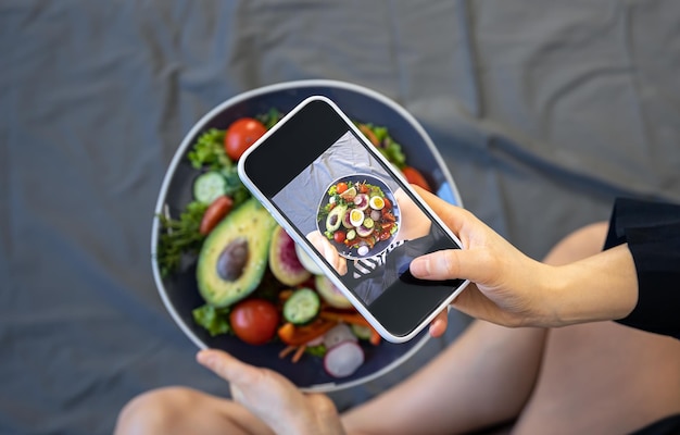 Una mujer toma una foto de una ensalada de vegetales frescos en un teléfono inteligente