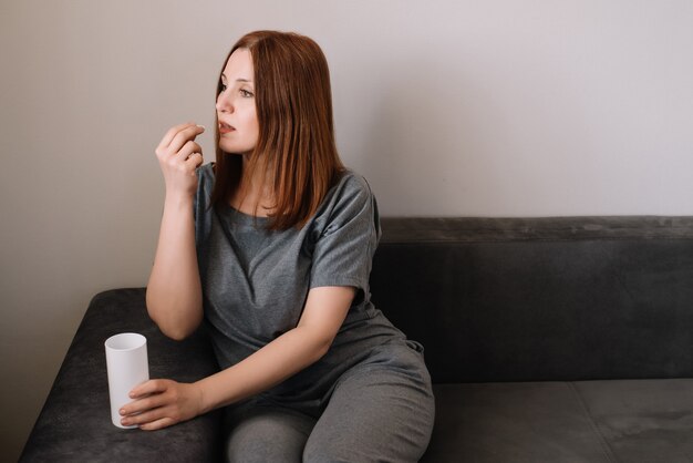 La mujer toma una píldora de vitaminas sentado en un sofá en la sala de estar en casa. Problema de coronavirus y concepto de protección covid-19.