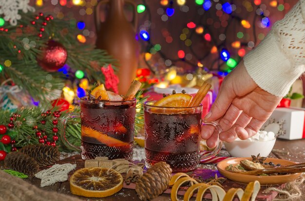 Una mujer toma una taza de vino caliente con una rodaja de naranja con el telón de fondo de las guirnaldas de luces festivas. Estilo oscuro