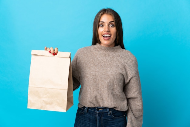Mujer tomando una bolsa de comida para llevar aislada en la pared azul con expresión facial sorpresa