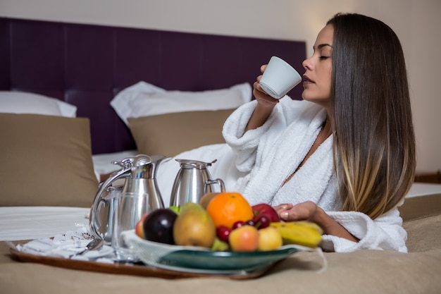 Mujer tomando café en la cama cerca de un plato de frutas