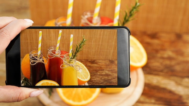 Mujer tomando foto de jugo de frutas en su teléfono inteligente. La mano sostiene un teléfono con una foto. Jugos de frutas en una brillante foto de verano. Sin marcas y logotipos visibles