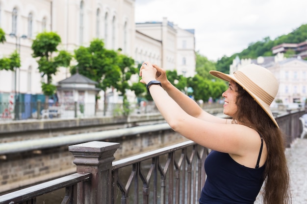 Mujer tomando fotografías con el teléfono inteligente. Mujer de viajero de verano con estilo en sombrero con cámara al aire libre en la ciudad europea, casco antiguo de Karlovy Vary en el fondo, República Checa, Europa.