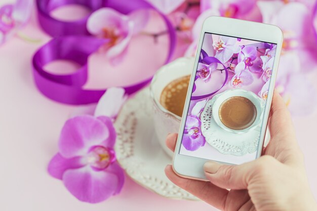 Mujer tomando una hermosa foto de una taza de café con orquídeas rosas y cintas púrpuras en la mesa con su teléfono inteligente