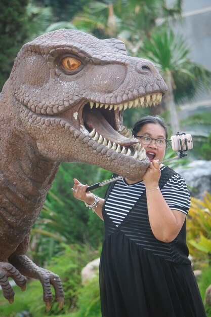 Foto mujer tomando una selfie con una estatua de dinosaurio en el parque