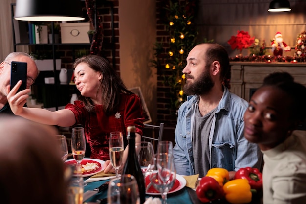 Foto mujer tomando selfie con smartphone en la cena festiva de navidad, sentada con la familia en la mesa. gente diversa feliz reuniéndose, celebrando la navidad, comiendo comida tradicional juntos