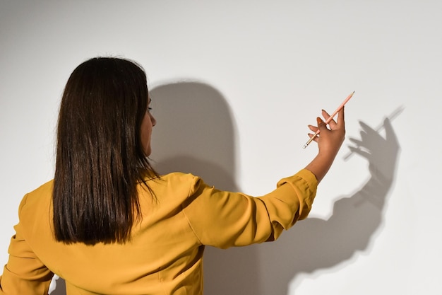 Una mujer con un top amarillo señala una pared blanca que tiene una sombra.