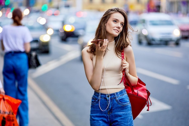 Mujer en top y jeans usa smartphone en la concurrida calle de la ciudad con tráfico de automóviles