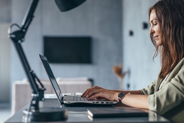 La mujer trabaja en casa sentada en su escritorio con la vista lateral de su computadora portátil