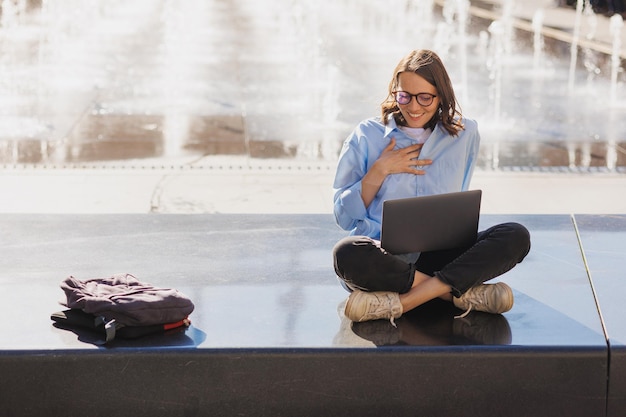 La mujer trabaja en una computadora portátil al aire libre en una persona de negocios de verano sentada junto a una fuente