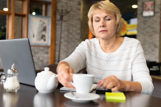 Una mujer trabaja con una computadora portátil en una mesa en un café o en la oficina. Una mujer de mediana edad trabaja de forma remota. Ella es una mujer de negocios.