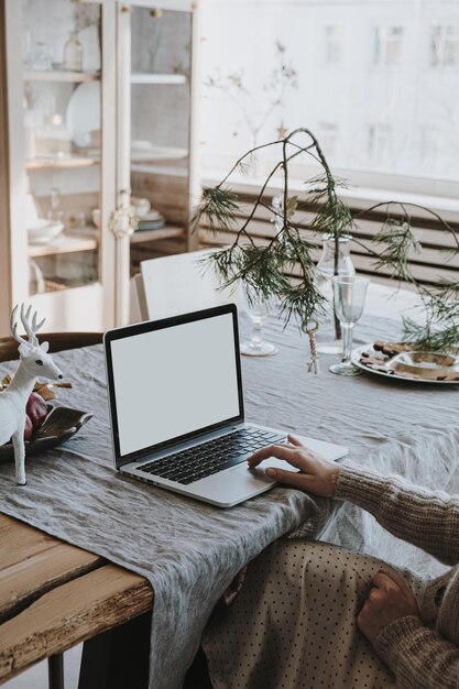 La mujer trabaja en una computadora portátil con una pantalla en blanco en un mantel de lino con un ramo de abetos Decoraciones navideñas Elegante oficina en el hogar espacio de trabajo diseño de interiores Freelancer trabaja en casa