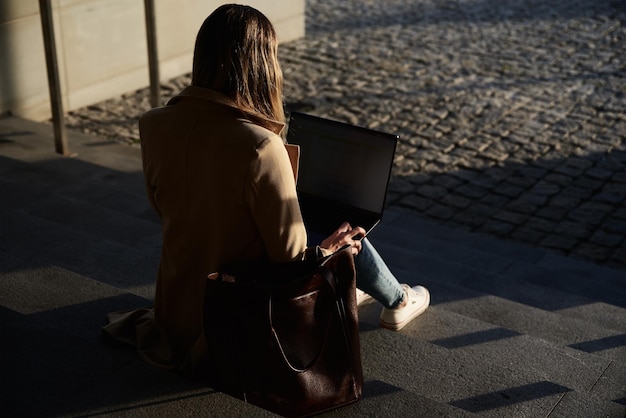 La mujer trabaja de forma remota en la computadora portátil de uso de la calle de la ciudad y se sienta en las escaleras.