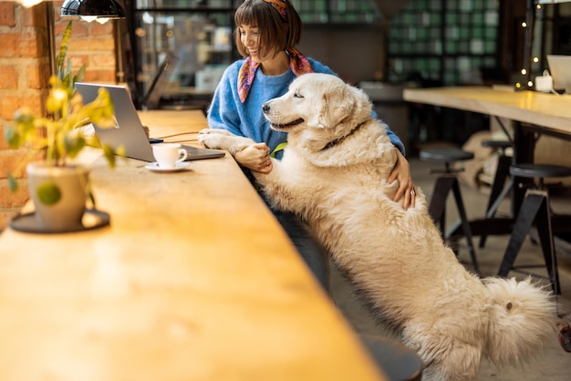 Una mujer trabaja en una laptop mientras se sienta con su perro en una cafetería moderna