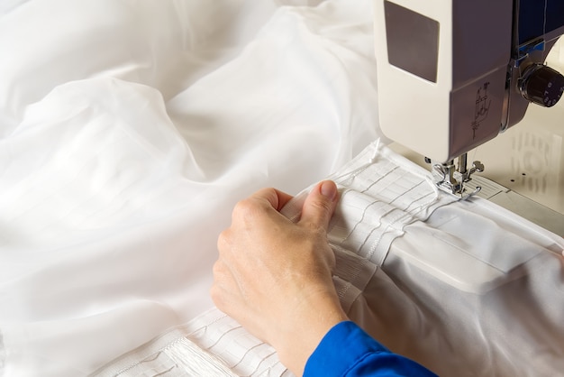 Una mujer trabaja en una máquina de coser. costurera cose cortinas blancas, vista cercana.