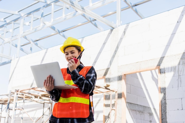 La mujer trabajadora arquitecta ingeniera asiática que trabaja en el sitio de construcción usa una computadora portátil y habla con la radio, la ingeniería sostiene la computadora y la radio discuten cómo operar y controlar a los empleados del edificio