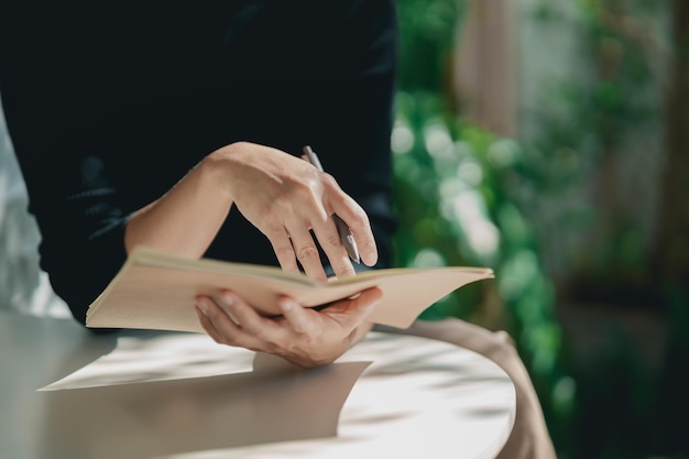 Mujer trabajadora asiática con camisa negra y escribiendo un diario en un pequeño cuaderno sobre la mesa en un café interior Notas de mujer y tomando café en el café Concepto de trabajo desde cualquier lugar