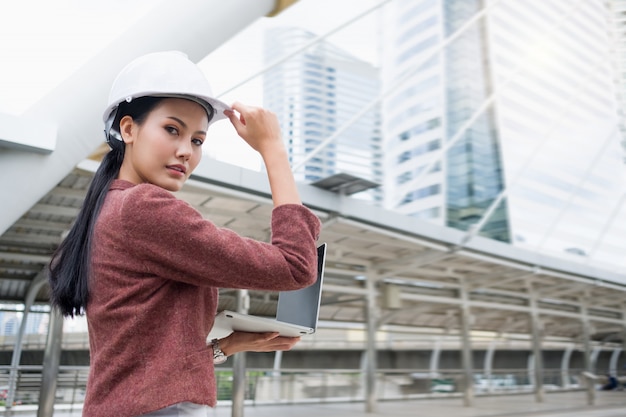 Foto una mujer trabajadora asiática confiada está usando un casco y trabajando en una computadora portátil mientras está de pie al aire libre.