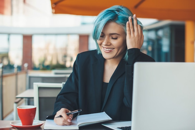 Mujer trabajadora con cabello azul escribiendo algo mientras está sentada en un restaurante y bebiendo un té cerca de su computadora