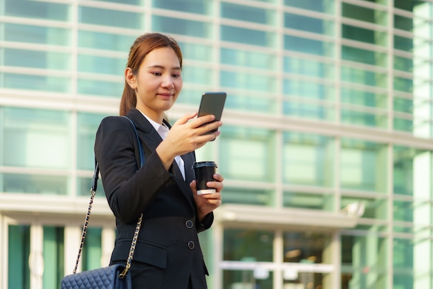 Mujer trabajadora ejecutiva asiática sosteniendo una taza de café y usando un teléfono móvil en la calle con edificios de oficinas en el fondo en Bangkok, Tailandia.