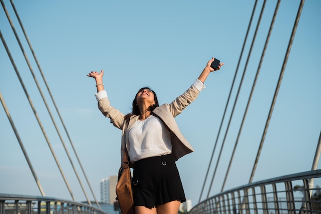 La mujer trabajadora levanta las manos con felicidad y triunfo en la calle sosteniendo un teléfono celular en la mano y está al aire libre Copiar espacio