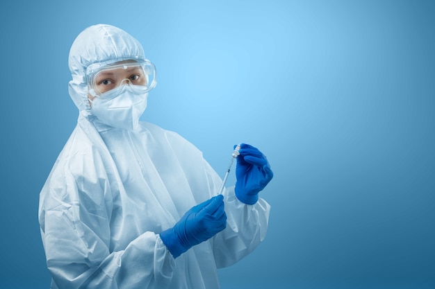 Mujer trabajadora de salud con traje protector y guantes sosteniendo vacunas covid 19 con fondo azul.