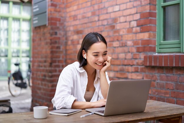 Mujer trabajadora sentada en un espacio de coworking tomando café y usando una computadora portátil con auriculares inalámbricos