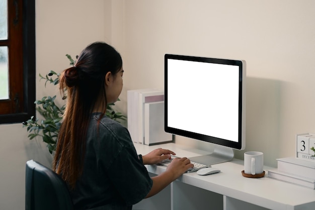 Mujer trabajando en una computadora de escritorio con pantalla en blanco en una oficina doméstica moderna y limpia