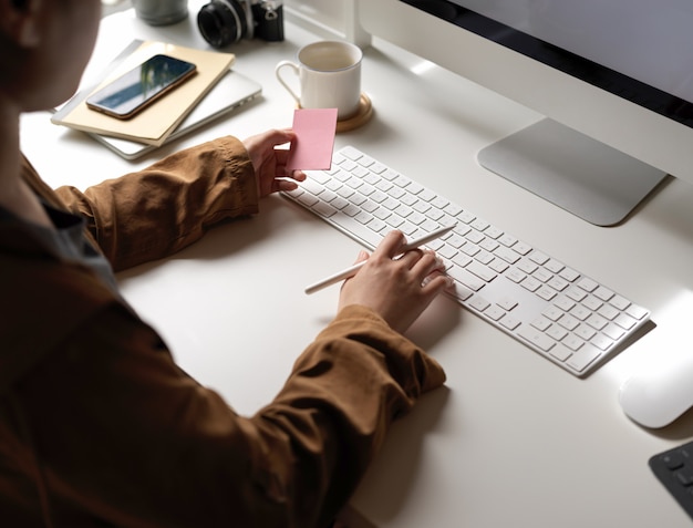 Mujer trabajando con dispositivo de computadora mientras busca una idea en el bloc de notas en el escritorio de oficina blanco