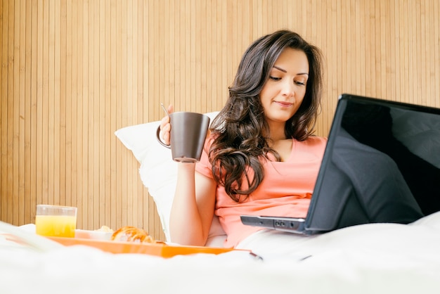 Mujer trabajando con su computadora portátil y desayunando. Ella está en su dormitorio.