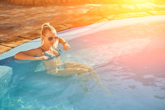 Mujer en traje de baño azul y gafas de sol relajante en la piscina al aire libre con agua turquesa transparente limpia ...