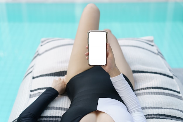 Mujer en traje de baño blanco y negro con teléfono móvil en blanco con pantalla en blanco junto a la piscina.