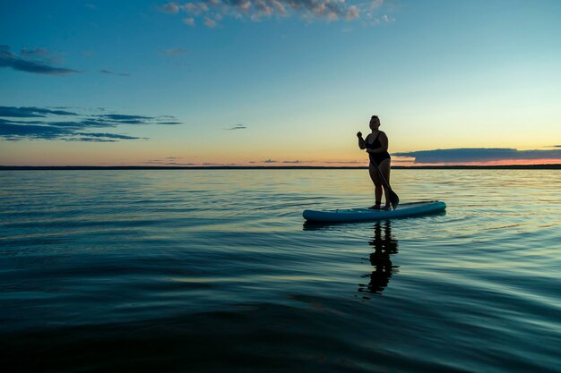 Una mujer en un traje de baño cerrado con un mohawk de pie sobre una tabla de SUP con un remo flota en el agua al atardecer