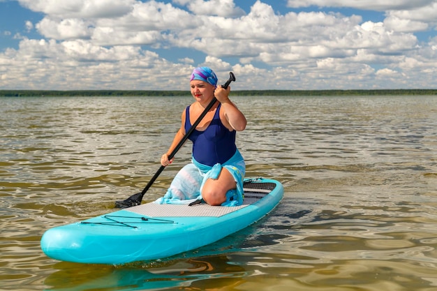 Una mujer con un traje de baño cerrado, un pareo y un tocado de rodillas en una tabla de SUP con flotadores de remo